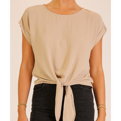 A woman wearing a beige short-sleeve top with a front knot detail, paired with black jeans, shown against a plain light beige background.